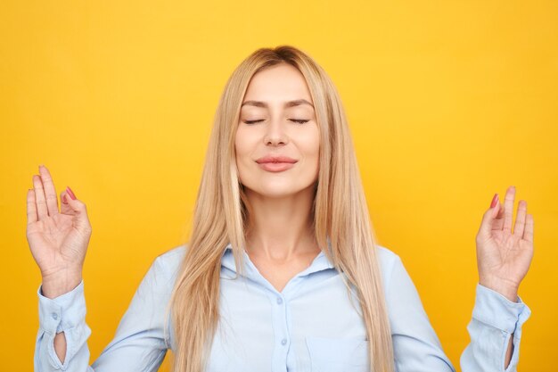 Businesswoman in a blue shirt keeps eyes closed holding fingers in mudra gesture, beautiful manager girl keeps calm relaxes isolated on yellow background, anti-stress therapy meditation