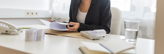 businesswoman consultant sitting at her office desk looking at paperwork and report, with receipts, notes, calculator and glass of water on her desk.