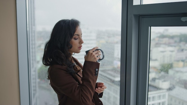 Businesswoman drinking coffee standing near office panoramic window close up