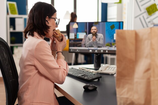 Donna d'affari che mangia panino da asporto durante la riunione di videochiamata online discutendo con un collega remoto. manager seduto alla scrivania con pranzo da asporto in ufficio