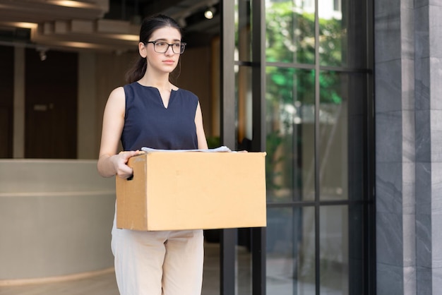 Businesswoman feeling sad carrying cardboard boxes walking from office after resign being fired