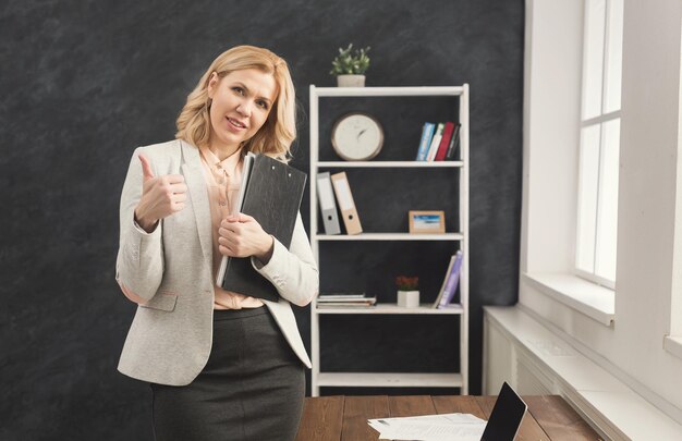 Businesswoman in formal wear holding documents and showing ok sign at modern office, copy space