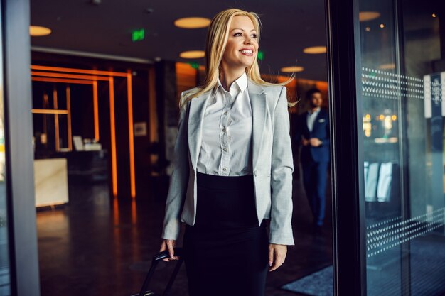 A businesswoman in a gray blazer, white shirt, and black skirt carrying luggage and leaving the hotel. Business trip, tourist destinations, ready for an official meeting