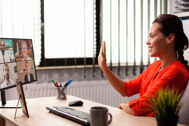 Photo businesswoman greeting employee with wave during video conference from workplace. entrepreneur using internet connection for video meeting with coworkers looking at webcam.