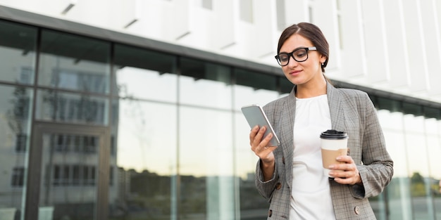 Photo businesswoman having coffee outdoors while looking at smartphone