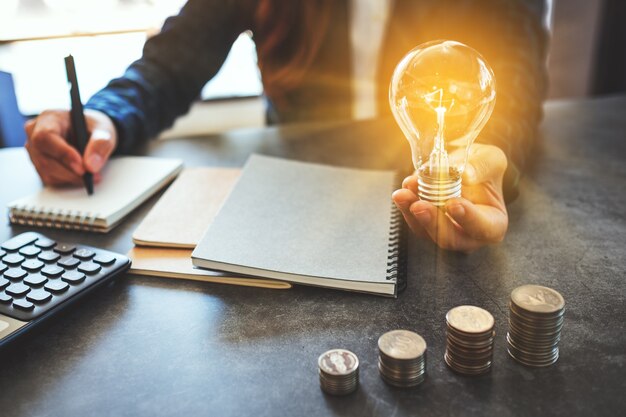 テーブルの上にコインを積み重ねてノートにメモを取りながら電球を保持し、エネルギーとお金の概念を節約する実業家