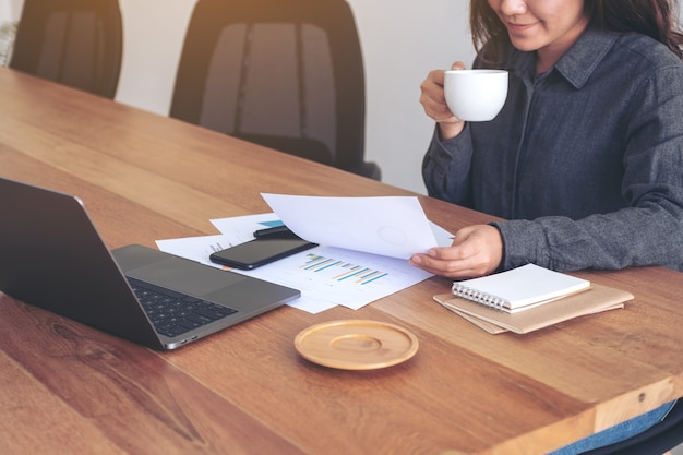 Una imprenditrice tenendo e guardando i dati aziendali e il documento con il computer portatile sul tavolo mentre beve il caffè in ufficio