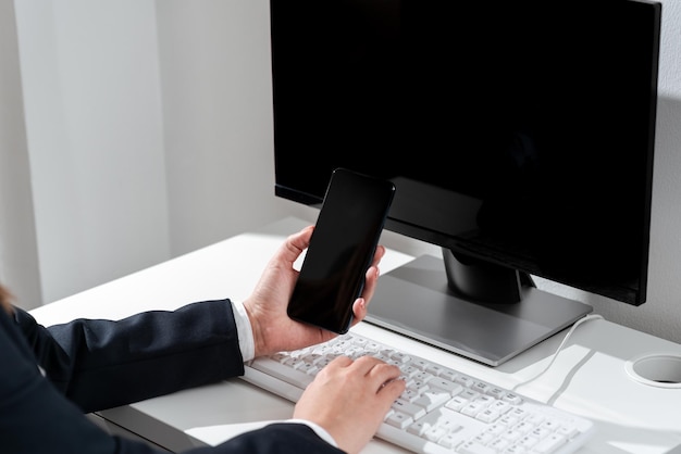 Деловая женщина держит мобильный телефон с важными сообщениями и печатает на компьютере на столе Женщина с мобильным телефоном в руке и записывает данные на ПК в офисе
