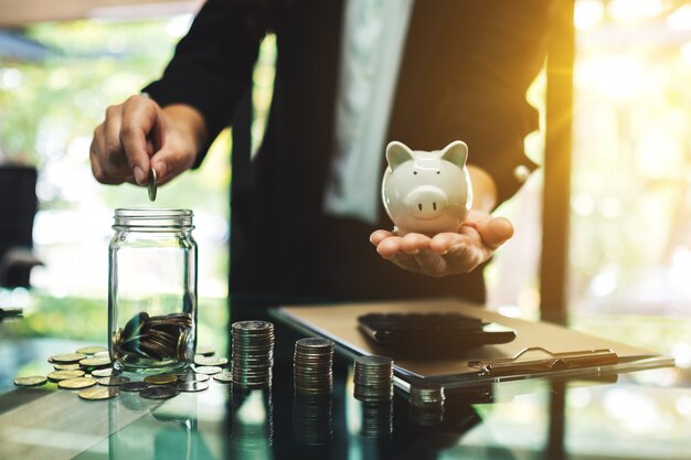 Businesswoman holding a piggy bank while putting coin into a glass jar for saving energy and money concept