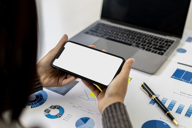 Businesswoman holding smartphone on desk with documents and laptop placed, she is looking at data from mobile phone, mobile phone blank screen for advertising graphics. the concept of using technology