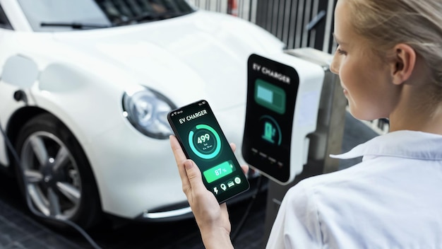 Businesswoman holding smartphone display battery status interface by smart EV mobile application while EV car recharging electricity from charging station in car park Peruse