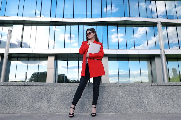 A businesswoman in a jacket poses with a laptop against the background of an office building