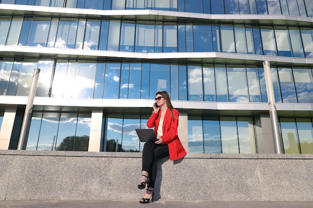 A businesswoman in a jacket works with a laptop and calls on the phone on the street