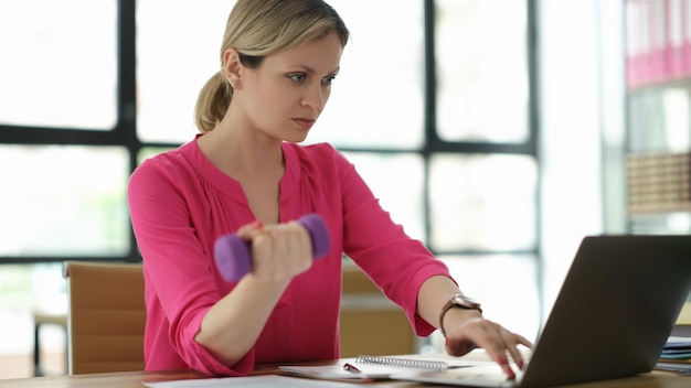 Donna di affari che solleva dumbbell e che lavora al computer portatile in ufficio