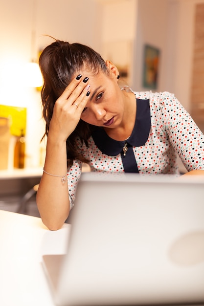 Businesswoman looking exhausted and massaging forhead while working overtime on laptop from home kitchen. Employee using modern technology at midnight doing overtime for job, business, career, network