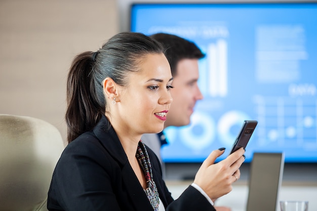 Businesswoman looking at phone in conferece room while a TV is displaying info in the background