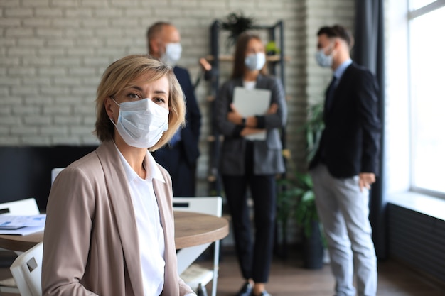 Businesswoman in medical mask with her staff, people group in background at modern bright office indoors.