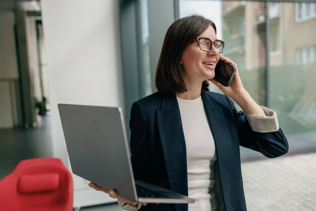 businesswoman multitasking with laptop and smartphone in modern office with glass windows