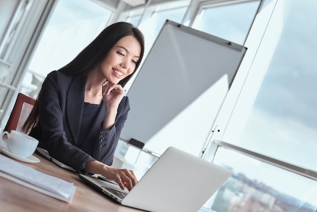 Деловая женщина в офисе одна сидит за столом с чашкой горячего кофе, просматривает Интернет на ноутбуке, улыбаясь счастливым