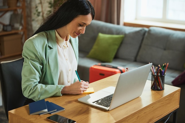 Businesswoman planning vacation sitting in her modern office and looking on laptop making notes