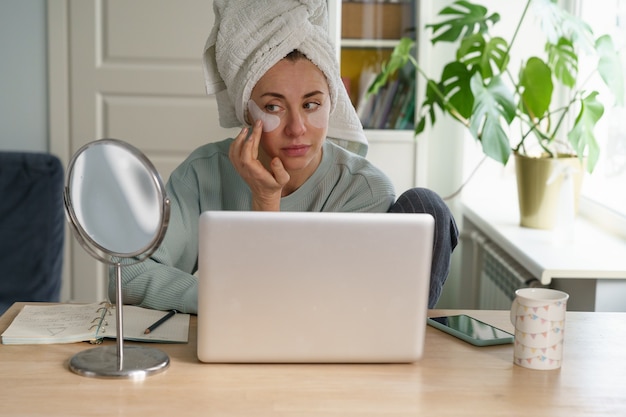 Businesswoman prepare for video conference on laptop with patches and with towel on hair in morning