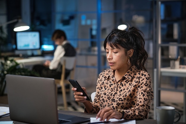 Businesswoman sitting at the table in front of laptop and reading a message on her mobile phone during her work at office