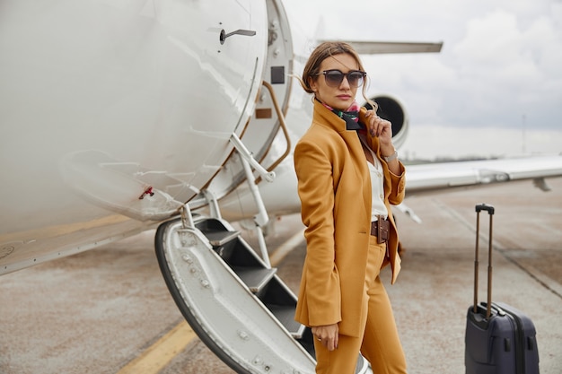 Businesswoman standing near private airplane jet. modern plane
with ladder. confident young woman wear formal suit and glasses.
civil commercial aviation. concept of air travel and business