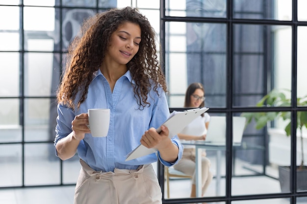 Businesswoman standing in the office corridor with documents and coffee Collegue is on the background