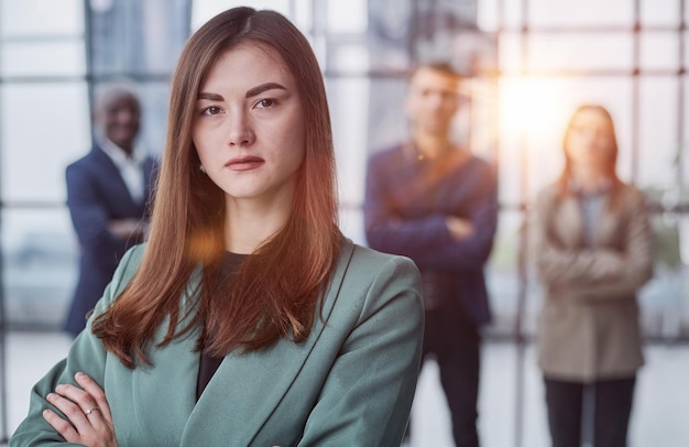 businesswoman standing with her arms crossed in an office while her colleagues have a meeting in the background