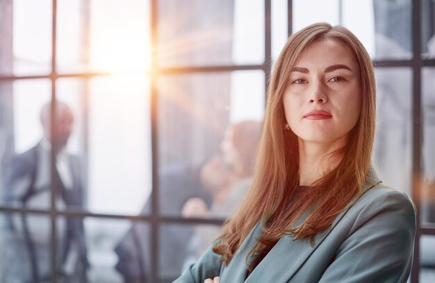 businesswoman standing with her arms crossed in an office while her colleagues have a meeting in the background