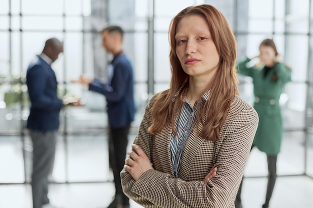 businesswoman standing with her arms crossed in an office while her colleagues have a meeting in the background