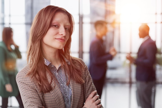 businesswoman standing with her arms crossed in an office while her colleagues have a meeting in the background