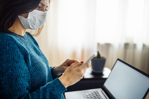 사진 사업가는 코로나바이러스 전염병 검역 기간 동안 집에 머물며 일합니다. 의료용 마스크를 쓰고 휴대전화를 손에 들고 있는 여성 노동자. 코비드-19 팬데미아 확산 예방 개념.
