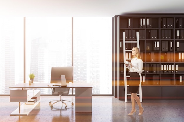 Businesswoman in stylish ceo office interior with gray walls, concrete floor, panoramic window, white and wooden computer table and gray bookcase with ladder. Toned image