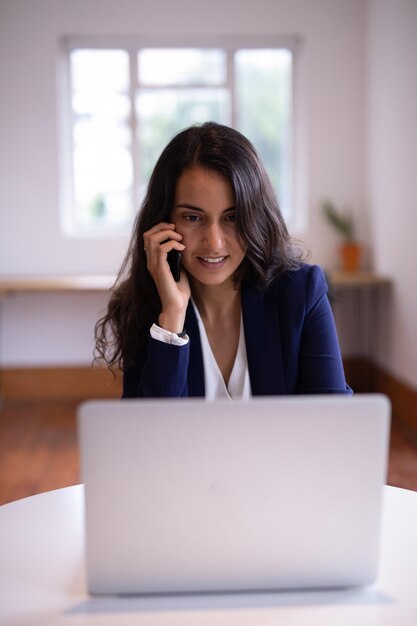 Businesswoman talking on mobile phone while working on laptop in a modern office