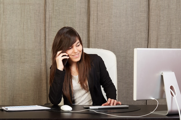 Donna di affari che parla con cellulare e che lavora nell'ufficio con il computer portatile