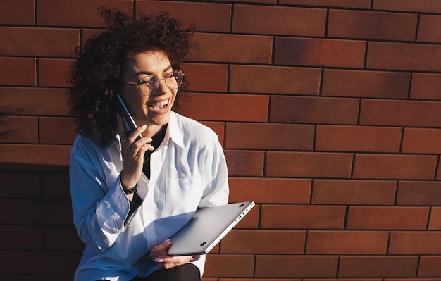 Businesswoman talking on the phone and holding a laptop in her hand while posing on a brick wall bac...