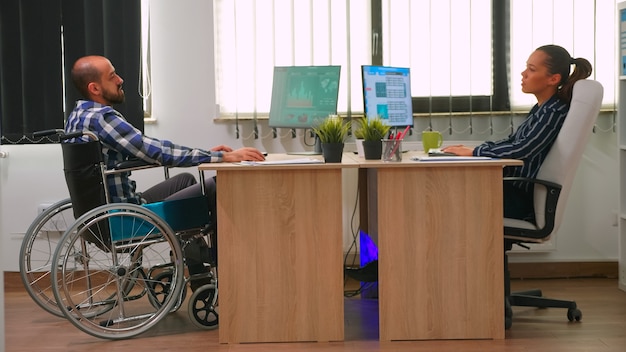 Businesswoman talking with paralysed colleague analyzing financial statistics, looking at desktop and typing. Disabled, immobilized freelancer working in corporate company using modern technology.