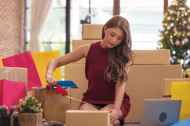 Businesswoman taping up a cardboard box, Asian woman packing boxes among stack of parcels in her shopping online business at home	