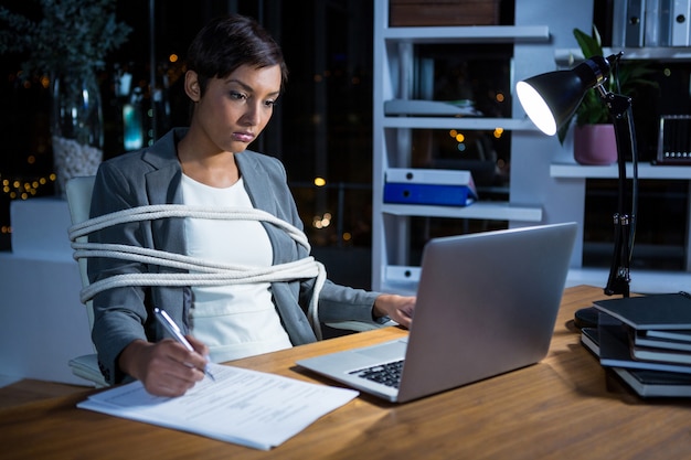 Businesswoman tied with rope while working on laptop at her desk