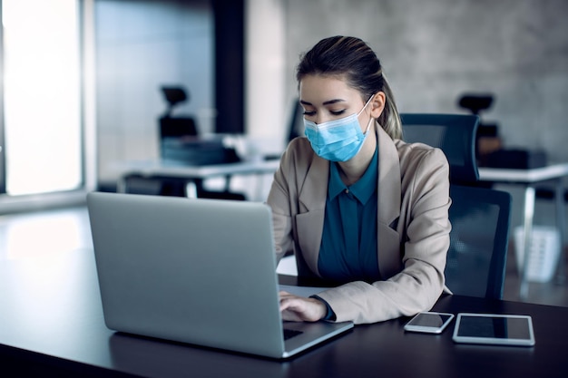 Businesswoman typing an email on laptop while wearing protective face mask in the office