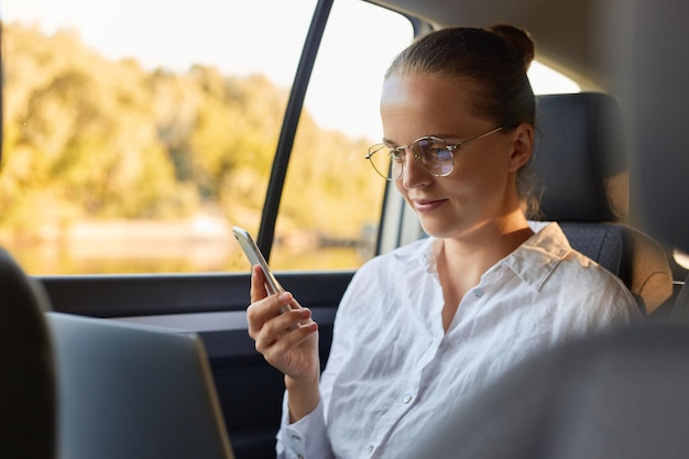 Foto imprenditrice utilizzando il telefono cellulare e il computer portatile in auto tutta seduta sul sedile posteriore comunicazione aziendale in movimento essendo concentrata sul display del notebook tenendo in mano il telefono cellulare