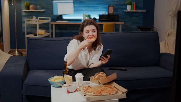 Businesswoman using smartphone while eating slice of pizza from delivery box on table. Young adult browsing on internet, having takeaway food for dinner in living room after work