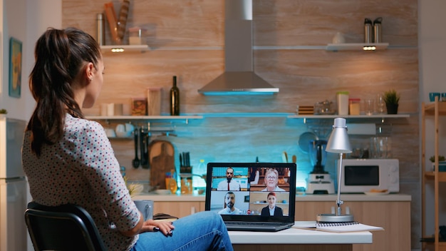 Businesswoman on video conference working remotely from home sitting in the kitchen late at night. Lady using modern technology network wireless talking on virtual meeting at midnight doing overtime