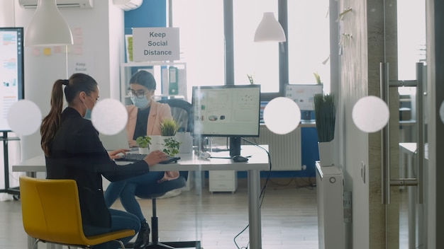 Donna d'affari che indossa una maschera protettiva che lavora al computer e discute con il collega sul lavoro d'affari. i colleghi mantengono il distanziamento sociale per evitare le malattie virali durante l'epidemia globale