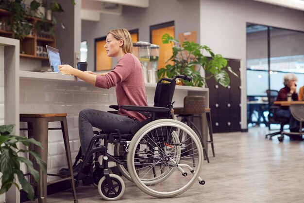 Imprenditrice in sedia a rotelle che lavora al computer portatile nella zona cucina di un ufficio moderno occupato