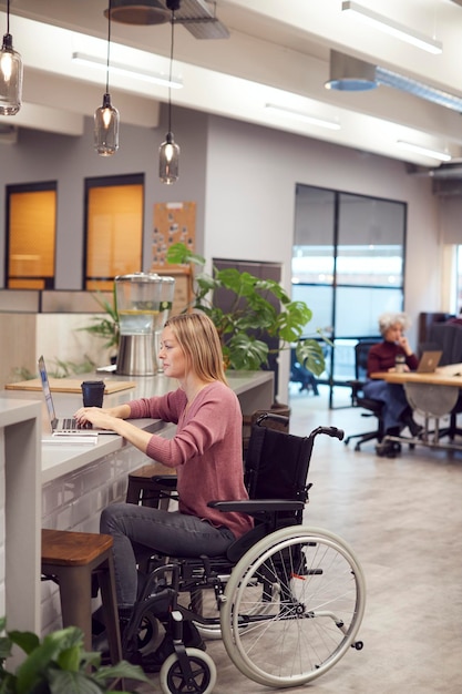 Foto imprenditrice in sedia a rotelle che lavora al computer portatile nella zona cucina di un ufficio moderno occupato