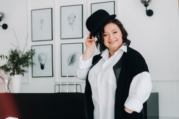 A businesswoman in a white shirt and trousers is standing with a hat in her hands standing in the office.