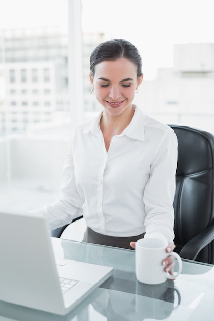 Donna di affari con la tazza di caffè mentre per mezzo del computer portatile allo scrittorio