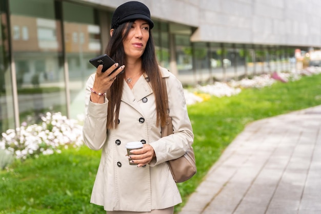 Imprenditrice con un caffè in mano e l'invio di una nota vocale con il telefono che indossa un trench